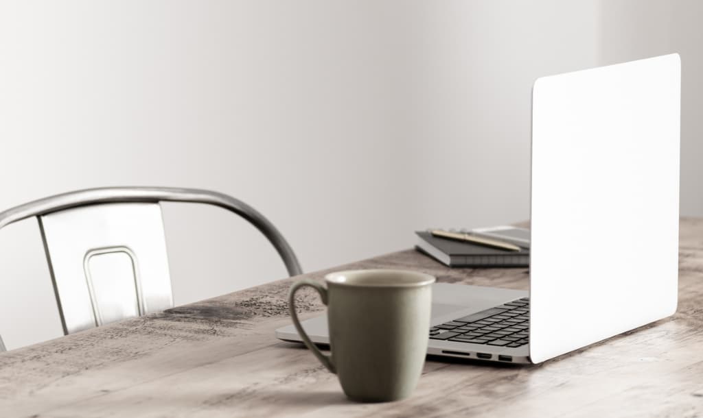 An open laptop and tan coffee mug sitting on a wooden desk with a tan metal chair sitting behind the desk.