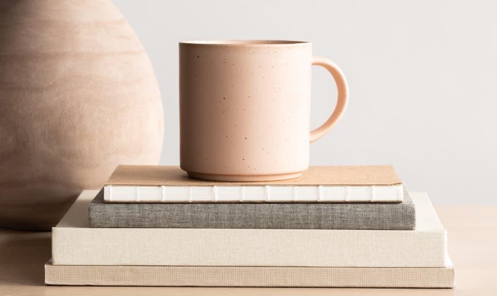 A blush colored mug sitting on top of a stack of books beside a blush coloured vase - How to Write Sleaze-Free Sales Page Copy