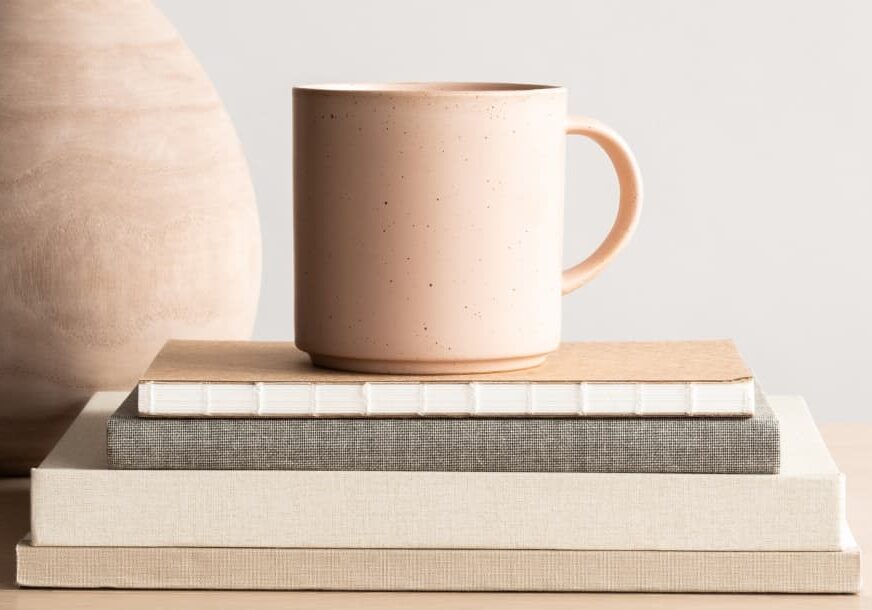 A blush colored mug sitting on top of a stack of books beside a blush coloured vase - How to Write Sleaze-Free Sales Page Copy